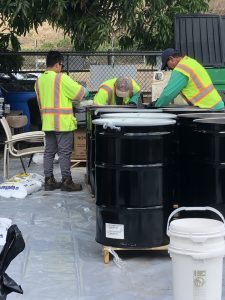 Three workers wearing high-visibility safety vests and gloves sort and handle materials near large black hazardous waste drums at an outdoor pesticide disposal site. The ground is covered with protective plastic sheeting, and bags labeled "DISPOSAL" are stacked nearby. Trees and fencing are visible in the background, and the setting appears organized for hazardous material collection.